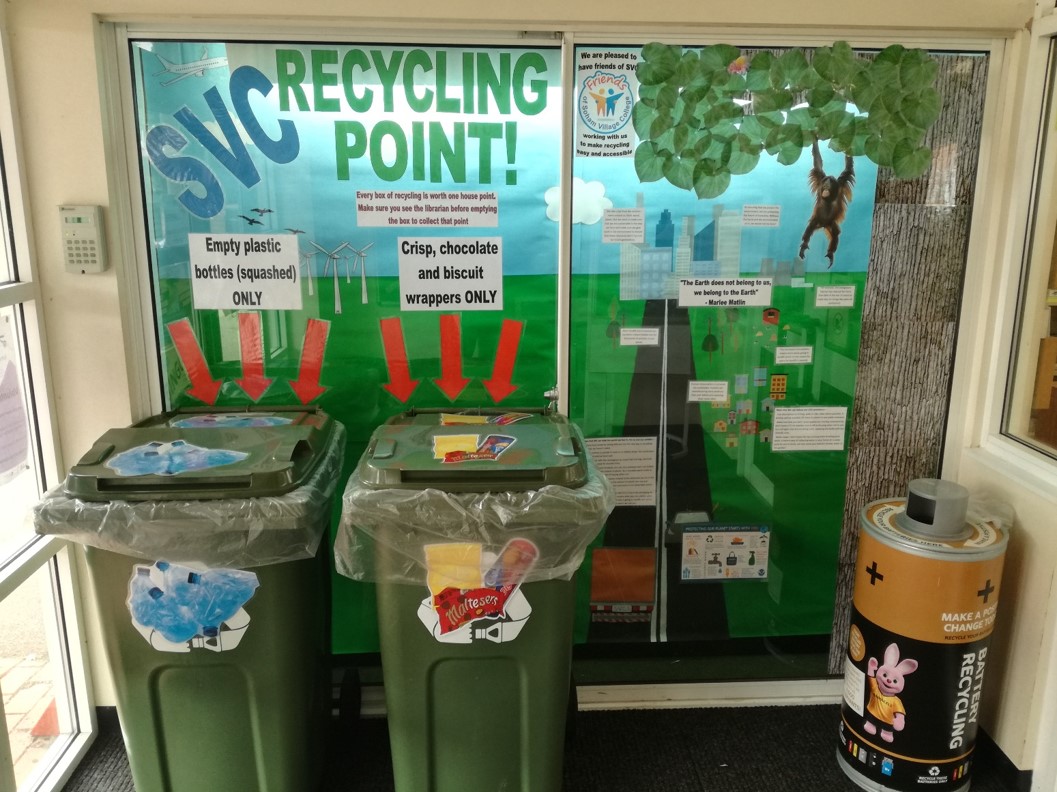 Picture of two green recycle bins, one for plastic and one for crisp and biscuit wrappers.  Recycle bin for batteries